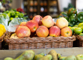 There is wicker basket with apples on counter in store. Container is filled to top with apples, and fruits are laid out in pyramid on showcase.