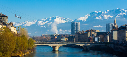 Cityscape of Grenoble with famous cable car against backdrop of snowy Alps in sunny day, France