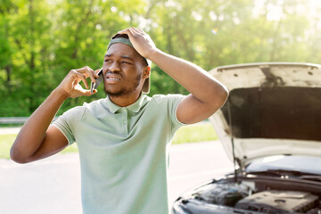 Unhappy black man standing near his broken car with open hood, experiencing engine failure, having...