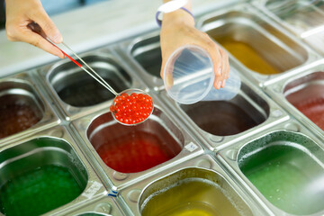 Hands of vendor preparing bubble tea, scooping red pearls from stainless steel container into plastic cup, surrounded by various colorful toppings on counter in tea shop