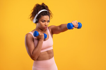 A young woman in a pink workout outfit lifts blue dumbbells while listening to music through headphones. The vibrant yellow background enhances her focused expression during the exercise.