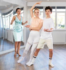 Motivated adolescent ballroom dancers, girl and boy in sportswear practicing elegant dance moves in pair in bright studio, with female coach supervising in background