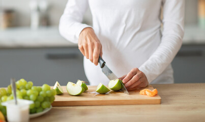 A pregnant woman is slicing green apples and tangerines on a wooden cutting board in a modern...