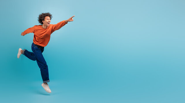 A young African American man with curly hair is energetically running and pointing at copy space, dressed in a casual orange sweatshirt and jeans.
