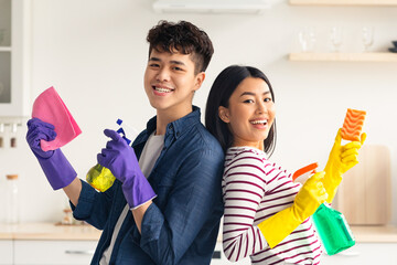 Cheerful asian couple with supplies for cleaning house posing in kitchen, funny young korean man...