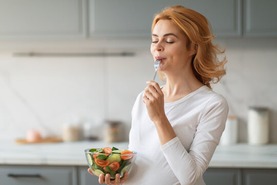 A young pregnant woman stands in a modern kitchen, savoring a bite of a colorful salad. She is smiling, indicating her enjoyment of the healthy meal amidst a clean, well-lit culinary space.