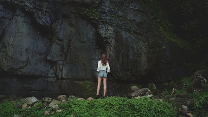 Young woman stands near the bottom of a jungle cliff face, taking in the natural beauty of a tropical island during the rainy season