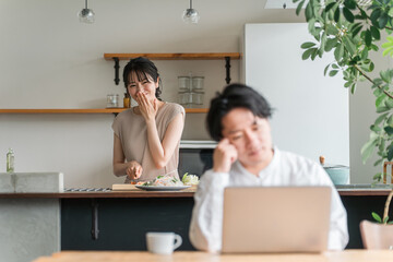 家でパソコン・テレワーク・リモートワークして悩む男性と料理する女性（夫婦）
