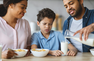 A father pours milk while a mother and son sit at the kitchen table. The son looks unsure about his food as they share a loving family breakfast together.
