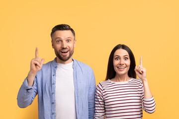 A happy couple stands together against a vibrant yellow background, both pointing upwards with joyful expressions. Their close bond and affection are evident as they celebrate a moment.