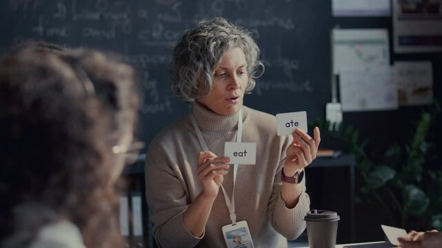 Middle-aged female teacher with curly gray hair holding flashcards and explaining verb tenses to students during English class
