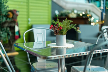 A festive natural Christmas centerpiece on a white doily with greenery, red berries, fir, pine cones, and buds. The arrangement is on a square silver metal restaurant table with four metal chairs. 