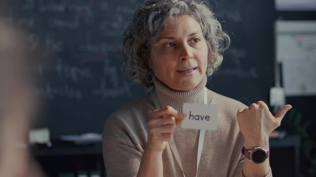 Middle-aged English teacher with curly grey hair showing flashcard labeled Have, explaining new word to students during lesson in language school