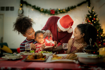 A joyful family gathers around the dinner table celebrating Christmas. The father, dressed in a Santa hat, gives a gift to the child, while everyone smiles with warmth and festive decorations.