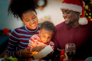A heartwarming Christmas moment as a young child opens a beautifully wrapped gift with the help of his parents. The mother gently guides the child while the father, dressed in a Santa hat.