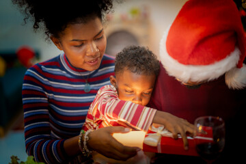 A heartwarming Christmas moment as a young child opens a beautifully wrapped gift with the help of his parents. The mother gently guides the child while the father, dressed in a Santa hat.