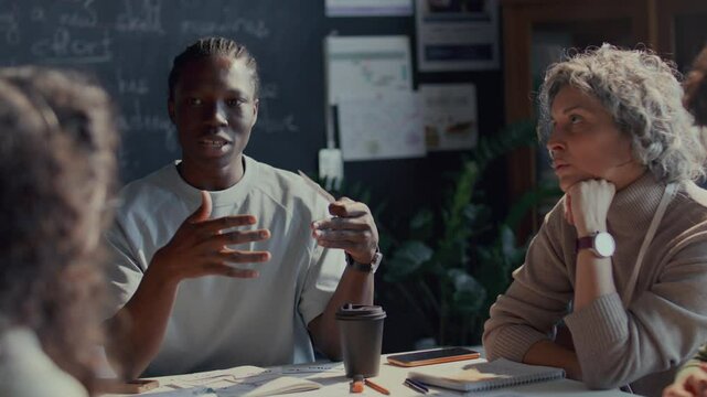 Black male student holding flashcard labeled Must, explaining its meaning during discussion with teacher and peers listens attentively during English class