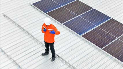 technician in hard hat and safety gear inspects solar panels on a metal rooftop, holding a tool....