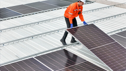technician in hard hat and safety gear inspects solar panels on a metal rooftop, holding a tool....