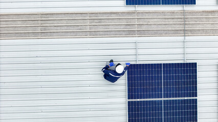 technician in hard hat and safety gear inspects solar panels on a metal rooftop, holding a tool....