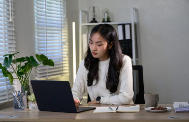 Young asian businesswoman is working on her laptop at her desk in a modern office with window blinds and plants, enjoying a cup of coffee and some cookies