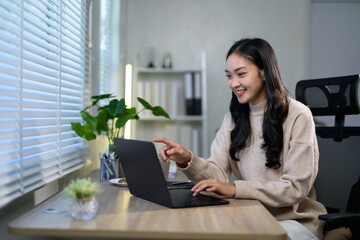 Young asian businesswoman working from home, sitting comfortably at a desk, smiling while using a laptop computer, with natural light streaming through the blinds of a nearby window