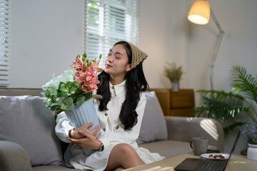 Young woman wearing a white dress is sitting on a sofa in her living room, smelling a bouquet of flowers in a blue vase, with a laptop and a cup of coffee on the table in front of her