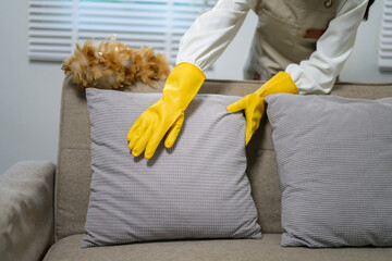 Housekeeper wearing yellow gloves is arranging pillows on a sofa in the living room, ensuring a...