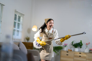 Young asian housekeeper wearing apron and gloves is mopping the floor in a modern living room, smiling and having fun while performing her chores