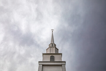 Church steeple with a cross on top rises against a dramatic, cloudy sky