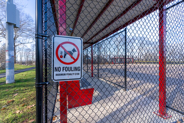 Close up photo of dog waste sign, No Fouling, hung on a black chain link fence at a baseball, softball field, park.
