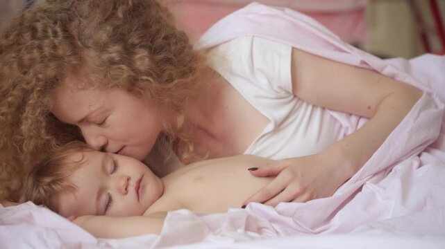 Mother with baby child lying on bed at bedroom. Beautiful mummy kissing and hug sleeping baby. Mother kissing sleeping baby. Mother caring for lovely child. Kid sleep in comfortable bed.