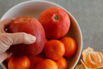 In a bowl, there are tangerines and apples. A hand is holding one apple. 