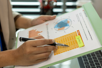 A woman is looking at a green clipboard with a lot of numbers and graphs on it. She is pointing at a specific number and seems to be analyzing the data. The clipboard is filled with various charts