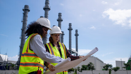 Engineers wearing safety gear, including hard hats examining survey a large blueprint tablet standing industrial facility gas or oil refinery engaged in a job requires high safety standards concept.