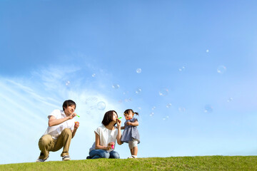 青空を背景にシャボン玉で遊ぶ若い夫婦と幼い娘。家族,親子,幸せ,愛情,育児のイメージ