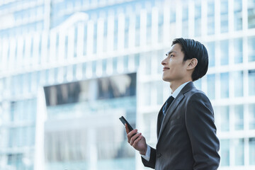 高層ビルのあるオフィス街で自身を持った表情でスマホを持ち遠くを眺める30代の男性