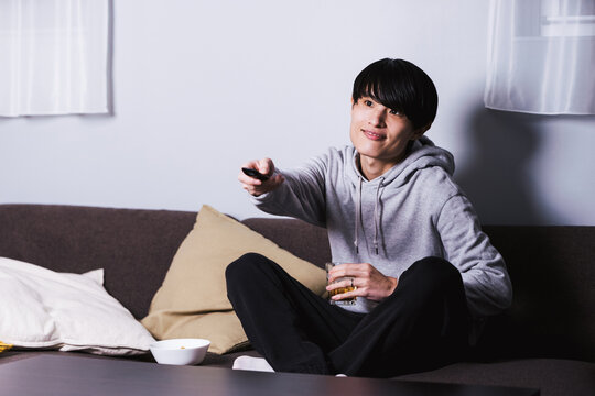 自宅の部屋で深夜にお酒とツマミを肴にテレビドラマやアニメを楽しむ為にリモコンを操作する30代の男性