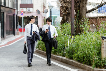 スマートフォンを見て会話しながら緑のある道路を並んで歩く若い2人のビジネスマンの男性