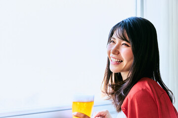 部屋の窓際で外を眺めながら美味しそうなビールを飲む30代のさわやかな笑顔の女性