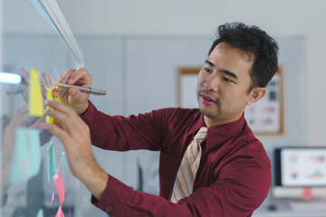 Businessman writing and pointing at sticky notes on glass to brainstorm and write on colorful note...