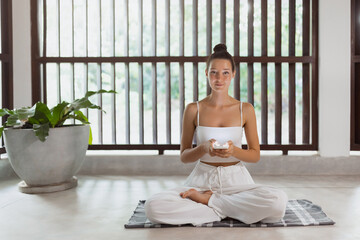 A calm woman in white sits cross-legged on a yoga mat, holding a smartphone. The bright, minimalist...