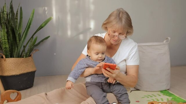 A grandmother and her young grandson sit comfortably on a floor mat. They share a joyful interaction as the grandmother helps the child explore the smartphone, promoting bonding and learning