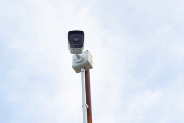 Surveillance camera mounted on pole against cloudy sky, capturing urban life. camera is positioned...
