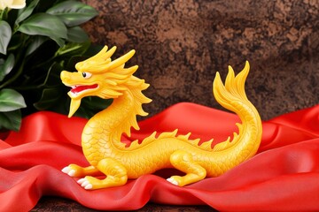 Fototapeta premium Chinese New Year symbolic gifts concept. A vibrant yellow dragon figurine rests on red fabric, surrounded by lush green leaves, creating a striking visual contrast.