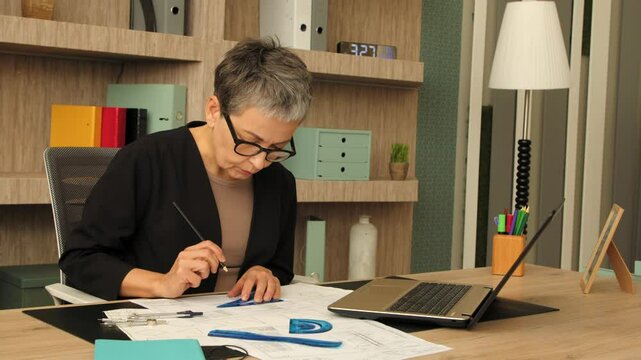 Concentrated professional lovely woman architect in eyeglasses working on new project , creating design and drawing blueprints using drafting tools at office .