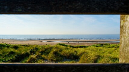Fototapeta premium The photo captures a stunning landscape of the Dutch coast in the Netherlands, featuring a wide sandy beach, lush green dunes, and the shimmering sea under a bright blue sky, inviting relaxation.