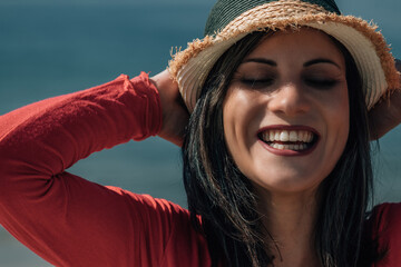 Photo of a Caucasian brunette woman in a straw hat smiling and sunbathing while walking on the beach