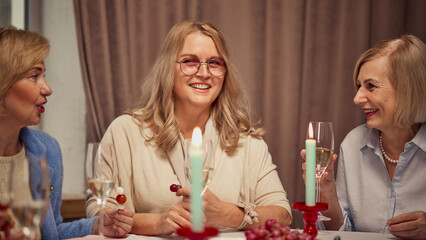 Happy laughing women on elegant clothes, sitting at table decorated with candles. in warmly lit living-room, drinking white wine. Concept of elderly lifestyle, positive emotions