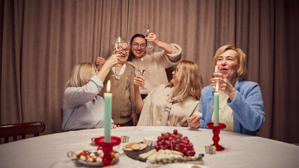 Happy laughing women, five friends cheerfully spending warm time at home, in warmly-lit living room, talking and drinking wine. Concept of elderly lifestyle, positive emotions, celebration